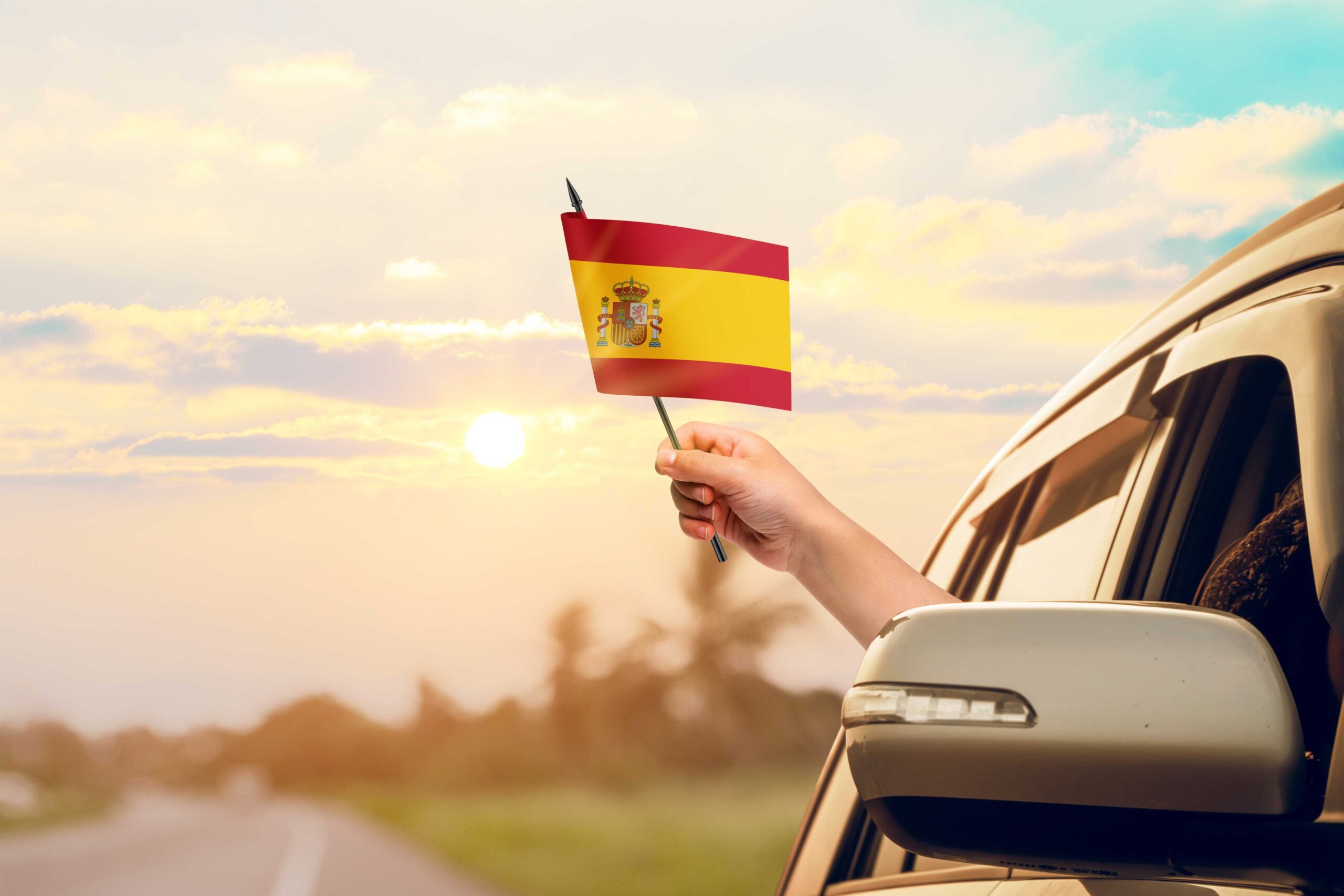 A hand waves a Spanish flag from a car window to highlight the importance of car rental excess insurance for people renting cars.
