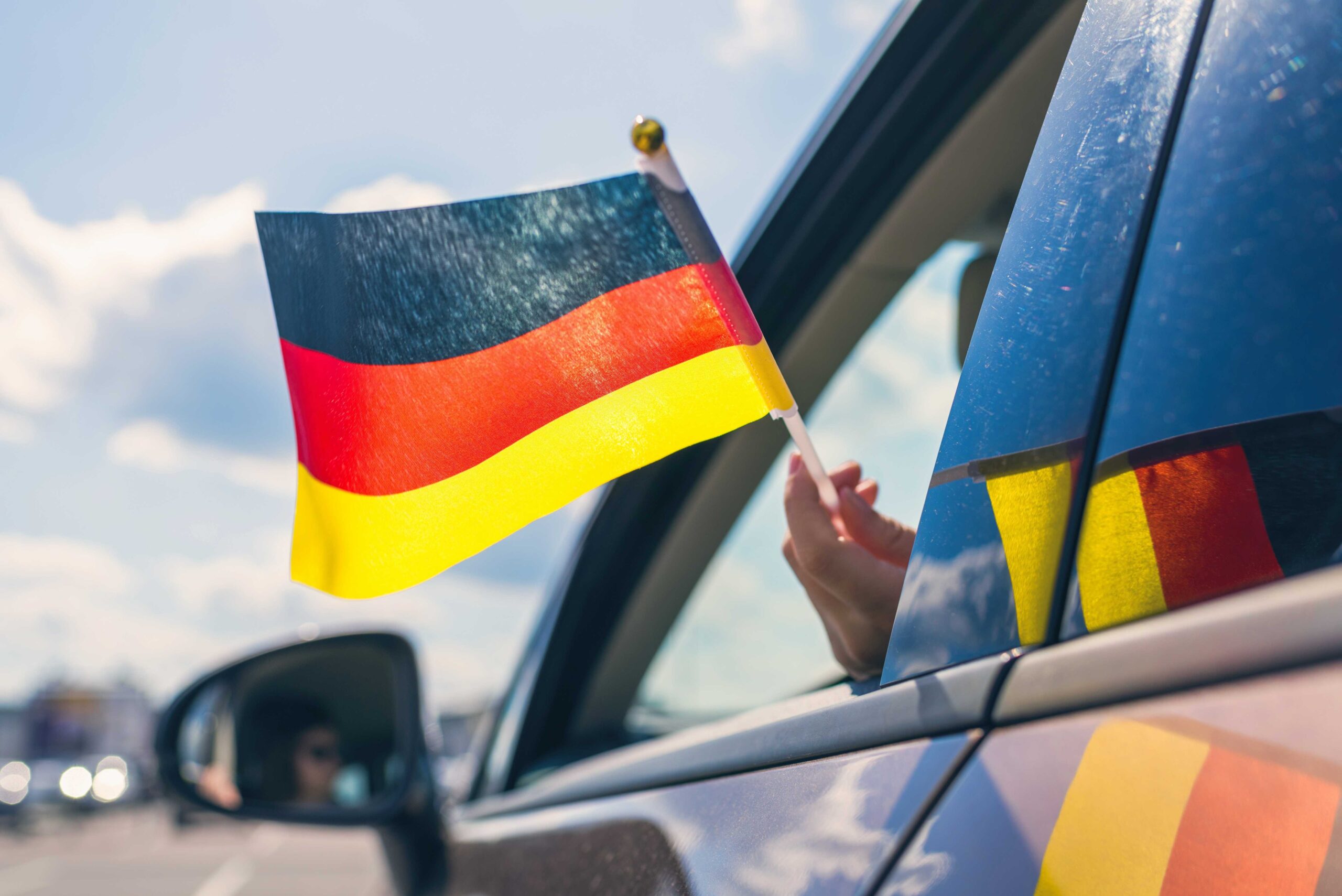 Person waving German flag outside of car window reinforcing importance of car rental excess insurance for people renting cars.