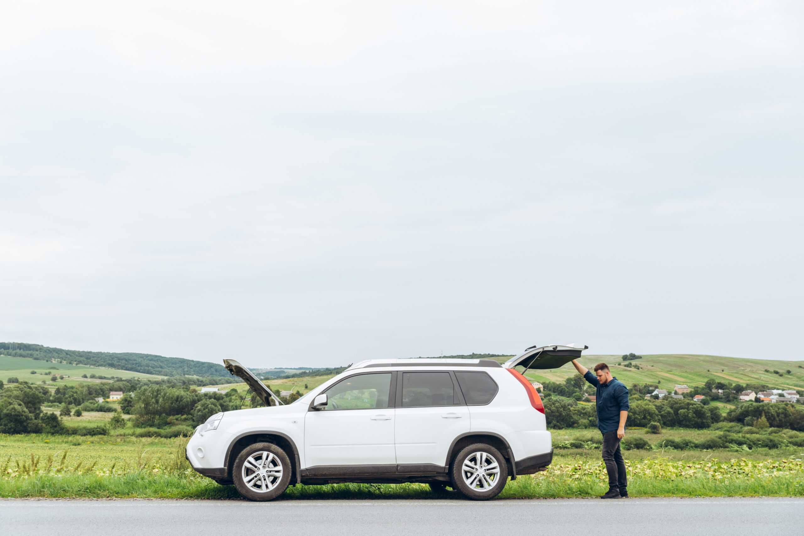 A white SUV is parked on a rural roadside with its hood and boot open.