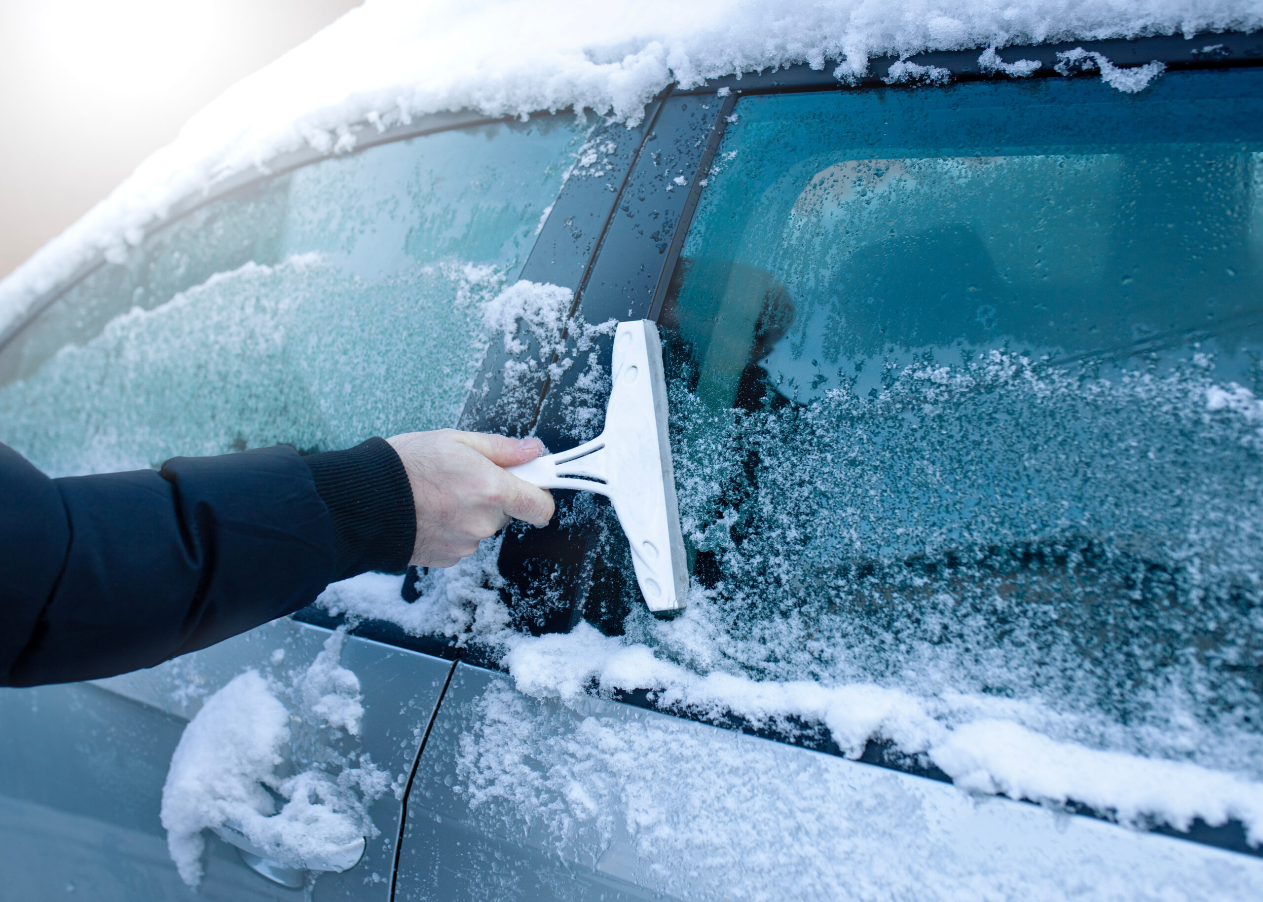 A hand uses an ice scraper to clear frost and ice from a car window.