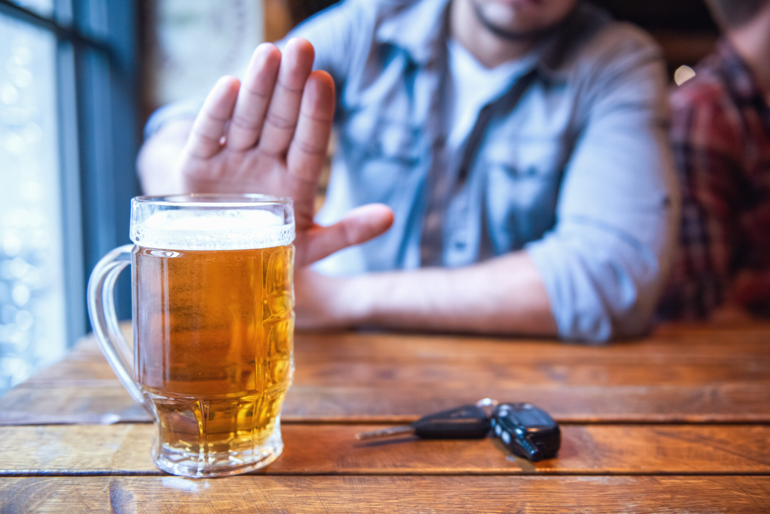 Person with hand raised in refusal gesture in front of a glass of beer on a wooden table with car keys nearby.
