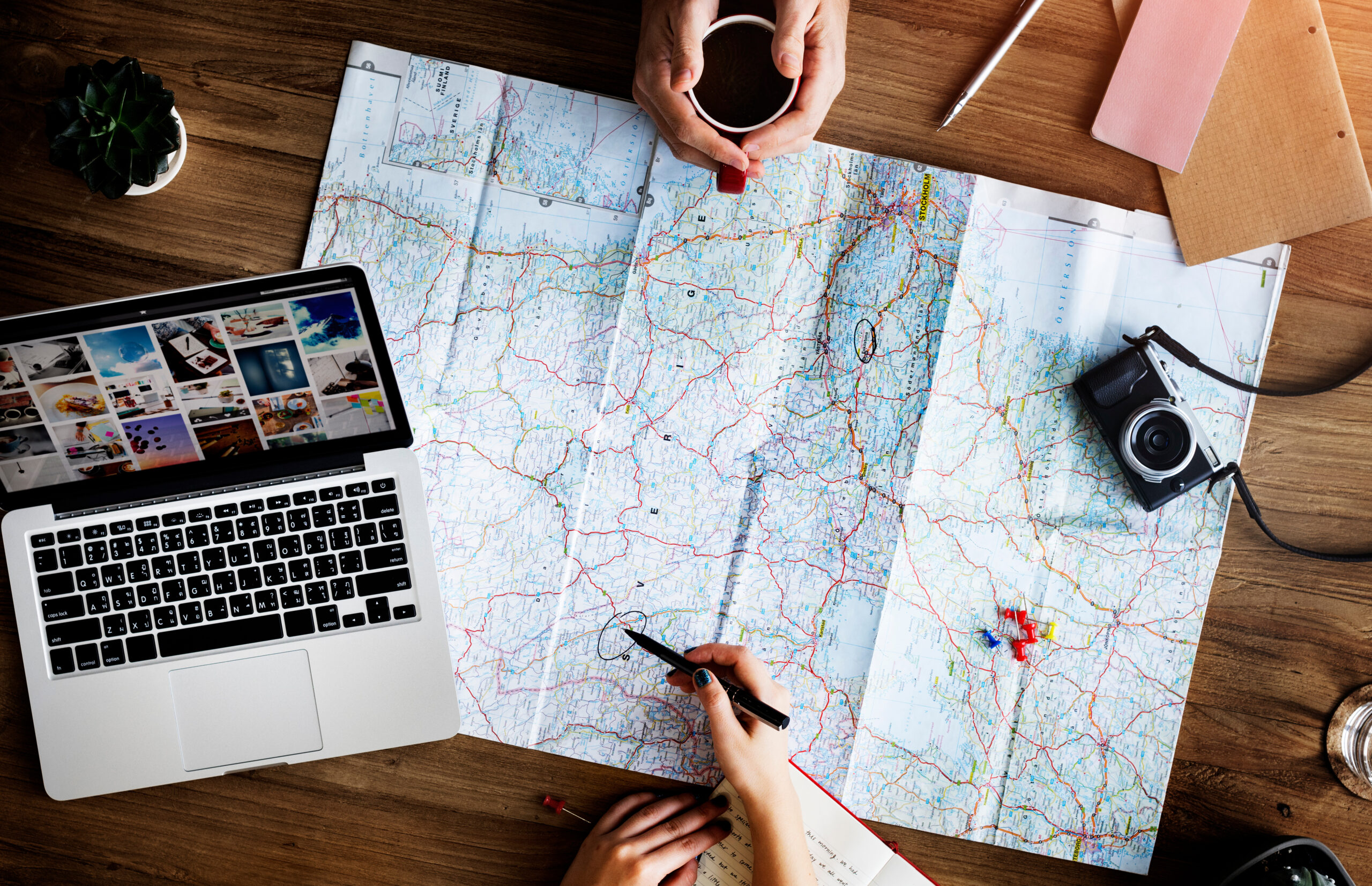 A wooden table with a spread-out map, a laptop displaying travel photos, a camera, and hands marking the map.